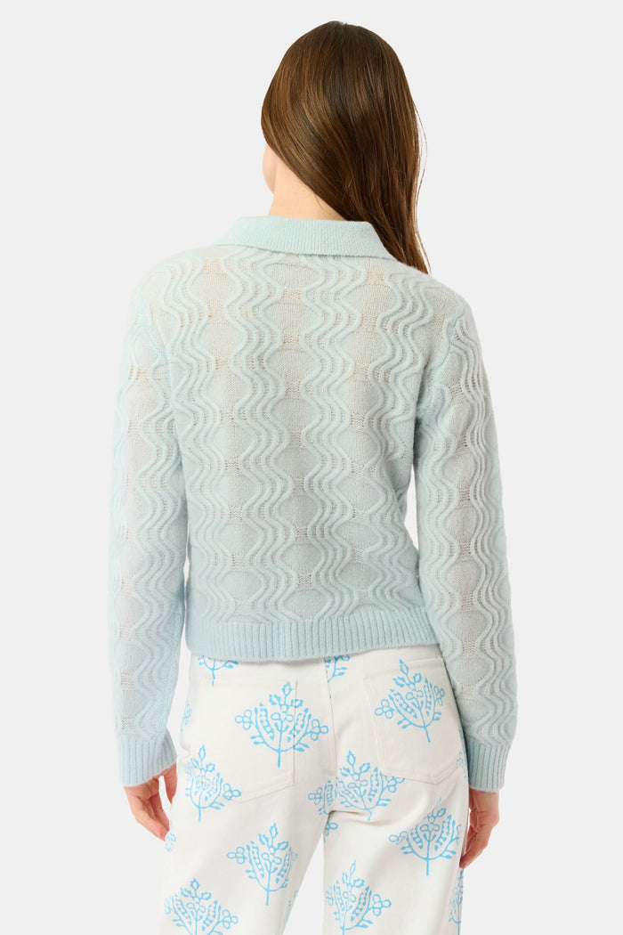 A woman with straight brown hair wears the Roller Rabbit Wave Knit Ava Sweater, a light blue collared, button-up style, paired with white pants with a blue floral print. She stands against a plain light background.