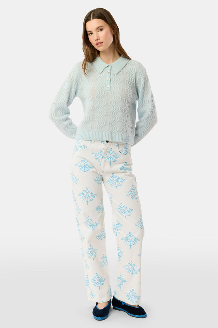 A woman with straight brown hair wears the Roller Rabbit Wave Knit Ava Sweater, a light blue collared, button-up style, paired with white pants with a blue floral print. She stands against a plain light background.