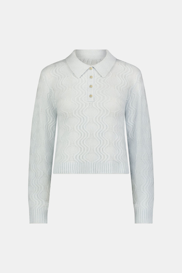 A woman with straight brown hair wears the Roller Rabbit Wave Knit Ava Sweater, a light blue collared, button-up style, paired with white pants with a blue floral print. She stands against a plain light background.