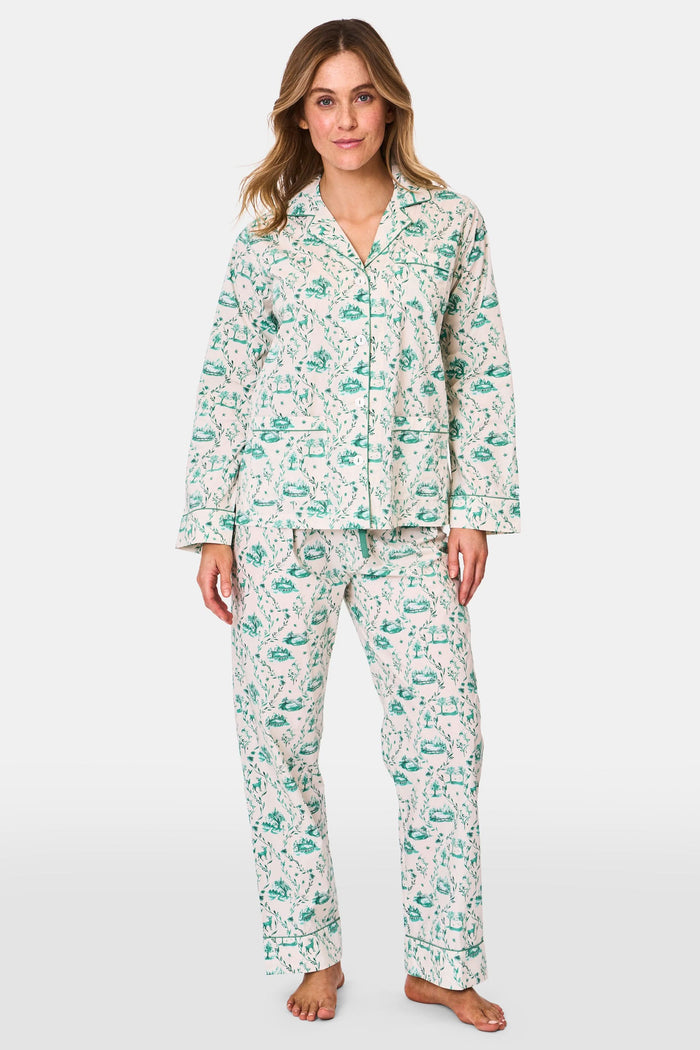 A woman stands barefoot in Roller Rabbit's Woodland Toile Loungewear, a cotton pajama set with a matching long-sleeve button-up top and pants in a green toile print. She faces forward, smiling slightly.
