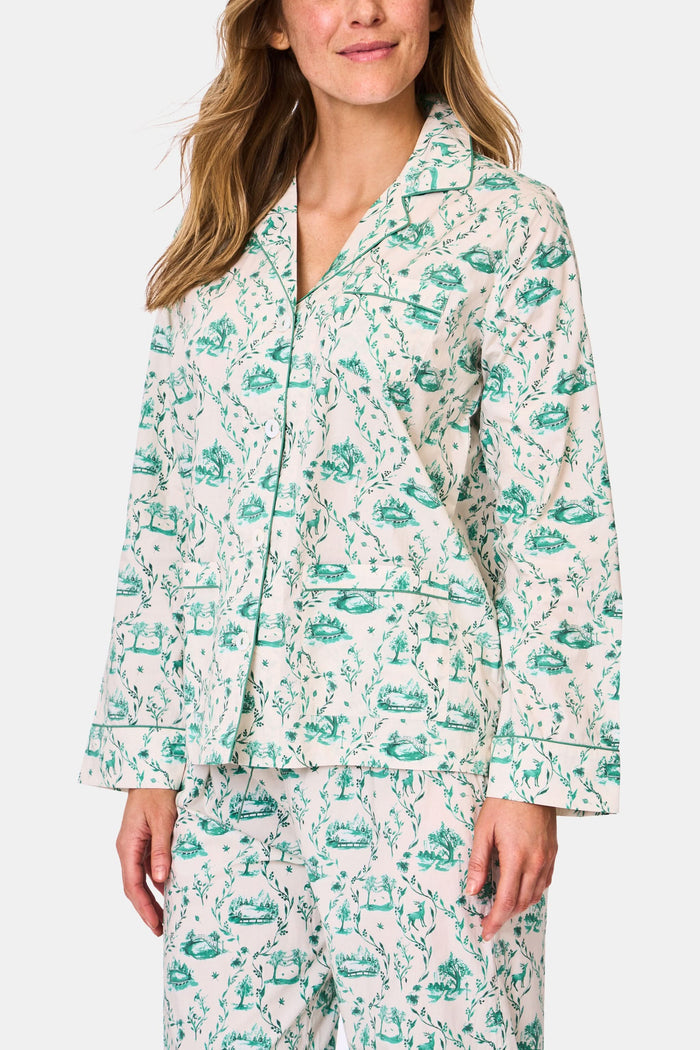 A woman stands barefoot in Roller Rabbit's Woodland Toile Loungewear, a cotton pajama set with a matching long-sleeve button-up top and pants in a green toile print. She faces forward, smiling slightly.
