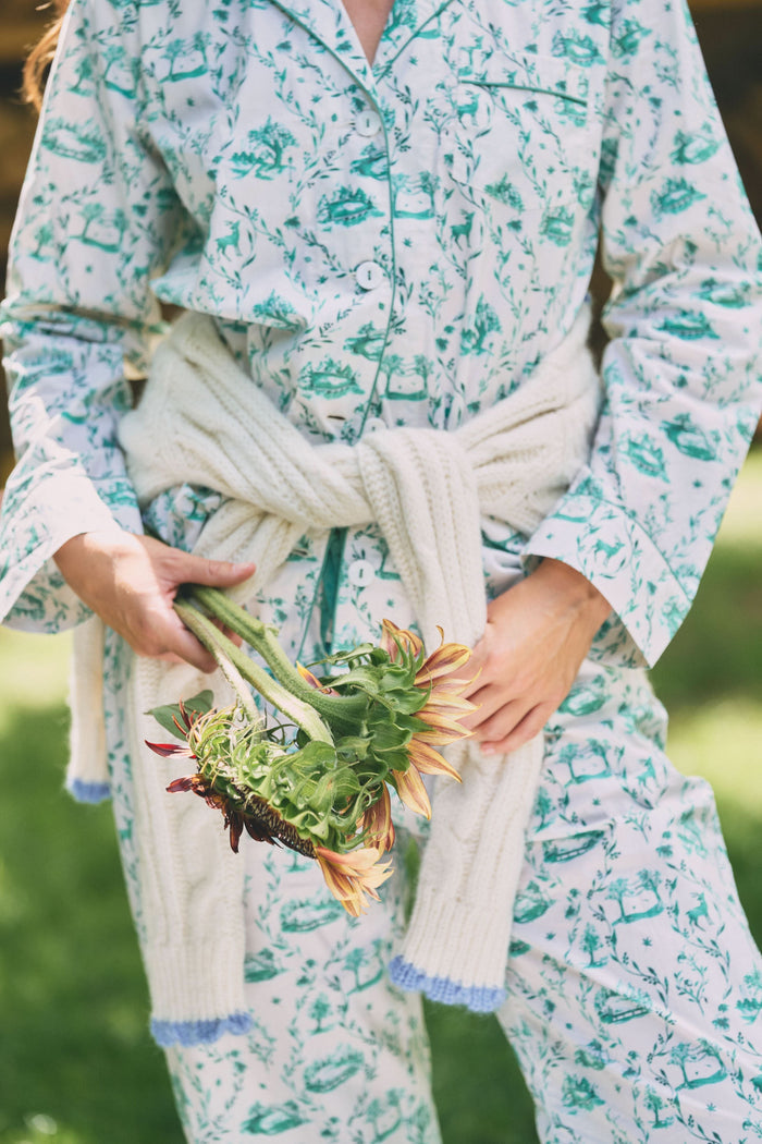 A woman stands barefoot in Roller Rabbit's Woodland Toile Loungewear, a cotton pajama set with a matching long-sleeve button-up top and pants in a green toile print. She faces forward, smiling slightly.