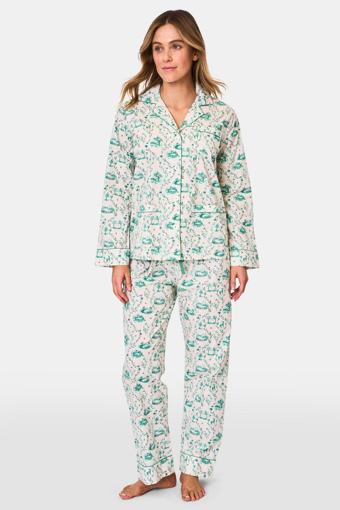 A woman stands barefoot in Roller Rabbit's Woodland Toile Loungewear, a cotton pajama set with a matching long-sleeve button-up top and pants in a green toile print. She faces forward, smiling slightly.