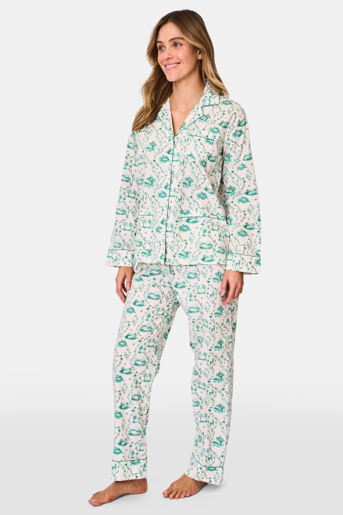 A woman stands barefoot in Roller Rabbit's Woodland Toile Loungewear, a cotton pajama set with a matching long-sleeve button-up top and pants in a green toile print. She faces forward, smiling slightly.