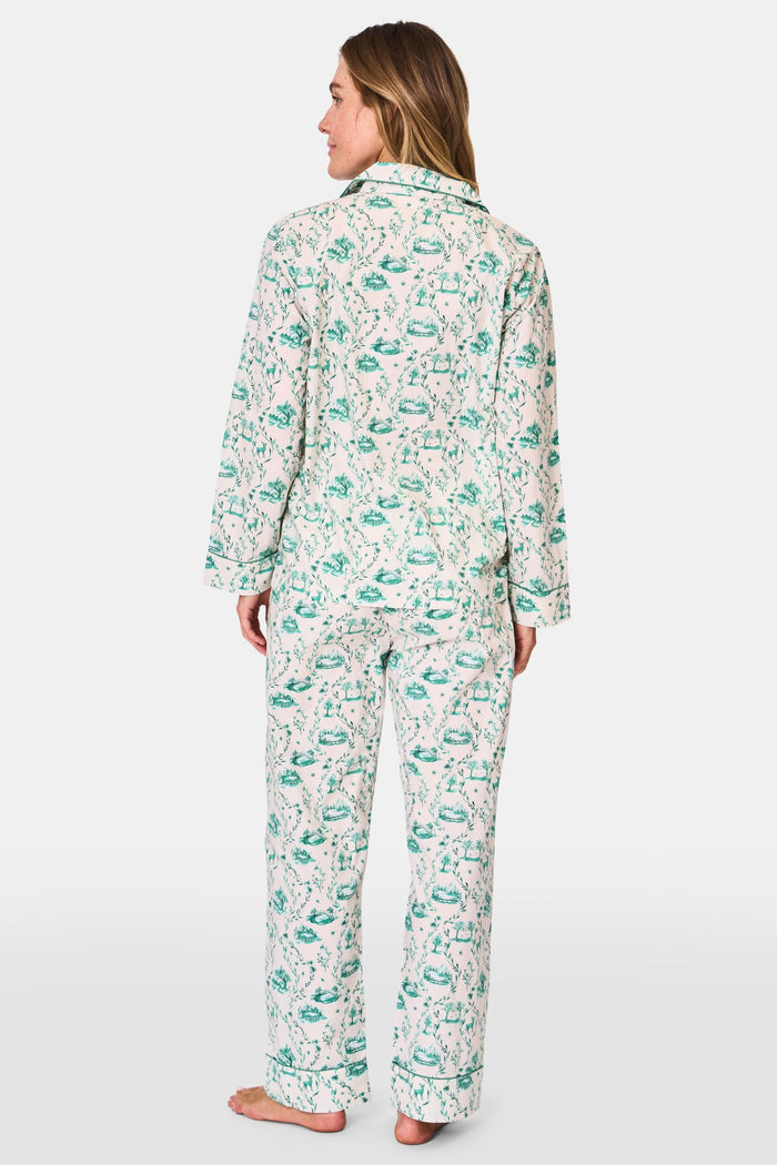 A woman stands barefoot in Roller Rabbit's Woodland Toile Loungewear, a cotton pajama set with a matching long-sleeve button-up top and pants in a green toile print. She faces forward, smiling slightly.