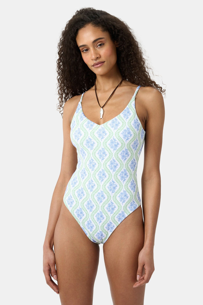 A woman with long curly hair wears the Roller Rabbit Zelda Floral Naia One Piece in blue, green, and white tones, standing against a light background. She looks at the camera with a calm expression and accessorizes her look with a necklace.