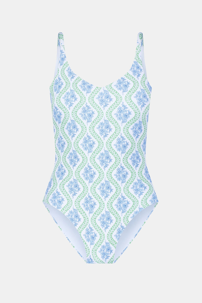 A woman with long curly hair wears the Roller Rabbit Zelda Floral Naia One Piece in blue, green, and white tones, standing against a light background. She looks at the camera with a calm expression and accessorizes her look with a necklace.